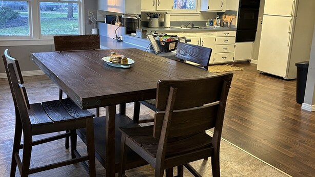A modern kitchen with white cabinets, a wooden dining table, and a view of the outdoors through large windows.