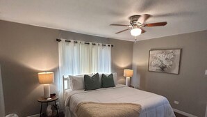 A bedroom with a bed, two bedside tables, a ceiling fan, a window with curtains, a rug, and a wall-mounted artwork.