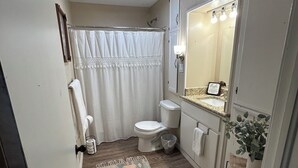 A bathroom with a toilet, shower curtain, rug, and a mirror with lights.