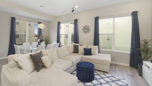 Una sala de estar moderna con un sofá seccional, un otoman redond, una mesa de comedor con sillas y ventanas grandes con cortinas.