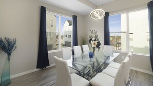 Un comedor con una mesa de vidrio, sillas blancas, un jarrón con flores y vistas a casas a través de amplios ventanales.