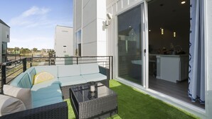 Un área exterior moderna con mobiliario, que incluye una mesa de vidrio, un sofá con cojines, y vistas al interior del edificio de al lado.