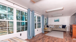 A spacious living room with large windows, a sectional sofa, a small stool, and a wooden floor. - Enjoy our speakeasy at our 2 bedroom cottage! (Sunrise Beach Village)