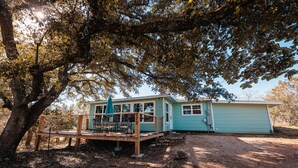 A small, light blue cabin with a wooden deck and outdoor seating area, surrounded by trees and a gravel driveway. - Enjoy our speakeasy at our 2 bedroom cottage! (Sunrise Beach Village)