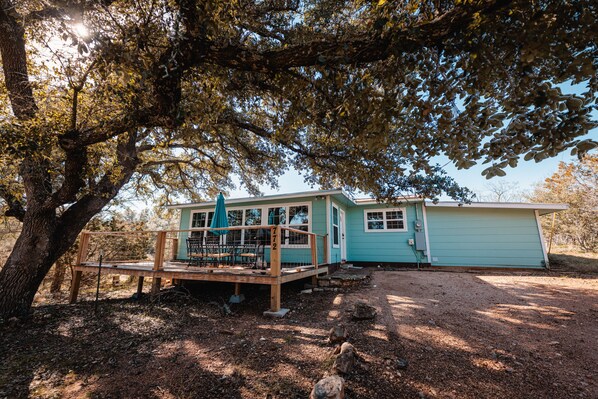 A small, light blue cabin with a wooden deck and outdoor seating area, surrounded by trees and a gravel driveway. - Enjoy our speakeasy at our 2 bedroom cottage! (Sunrise Beach Village)