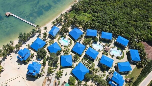An aerial view of a resort with multiple blue-roofed buildings, a beach, and a pier extending into the water.