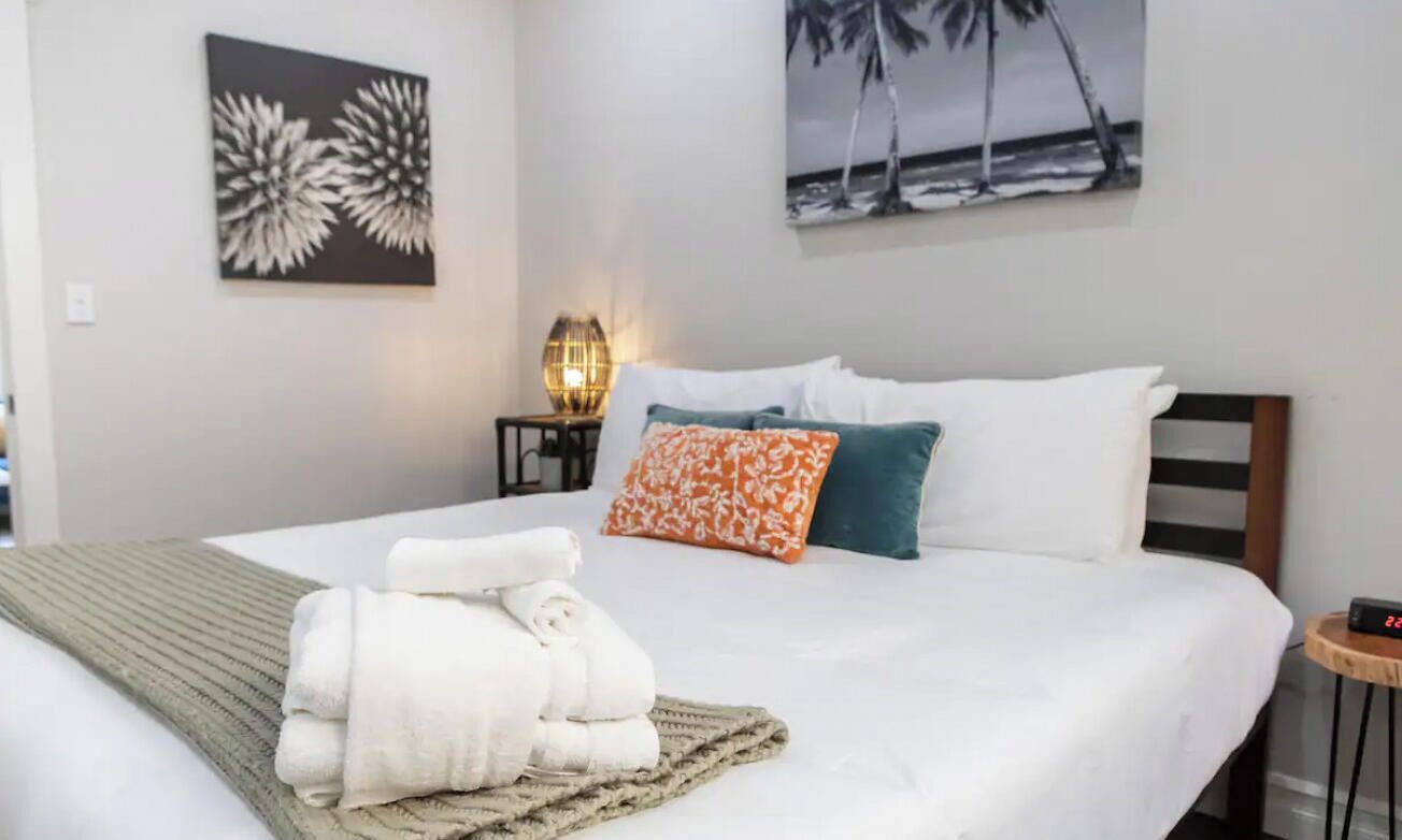 Un lit bien fait, avec du linge de lit blanc, une serviette pliée et un plaid décoratif. On y trouve une table de chevet avec une lampe et une horloge.