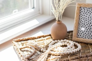 A wicker tray with a beaded necklace, a vase with dried plants, and a polka-dotted pillow on a windowsill.