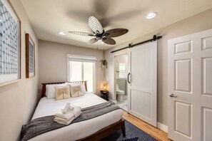 A bedroom with a bed, a ceiling fan, a bedside table with a lamp, and a bathroom with a toilet and sink.