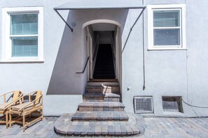 A building entrance with a set of stairs leading up to a door, a small outdoor seating area with wooden chairs, and a window with blinds.