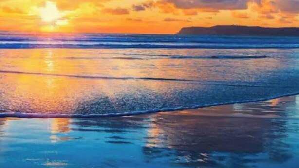 Una playa con un atardecer, agua tranquila y una masa de tierra a lo lejos.