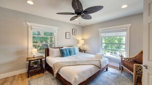 Un dormitorio amplio con una cama grande, dos mesitas de noche, un ventilador de techo y ventanas con vistas a zonas verdes.