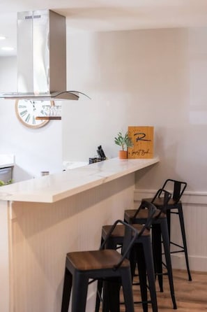 A modern kitchen with a white countertop, a clock, a potted plant, and two black bar stools.