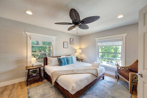 A bedroom with a bed, two bedside tables, a ceiling fan, and windows with views of greenery.