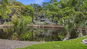Um edifício de dois andares com varanda, rodeado por vegetação exuberante e um lago tranquilo.