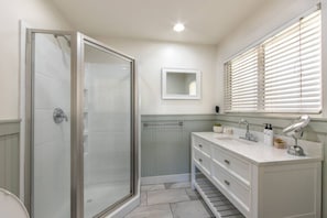 Un baño con cabina de ducha de vidrio, tocador con espejo y ventana con persianas.