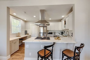 A modern kitchen with white cabinetry, a stainless steel range hood, and a central island with bar stools. - Coronado Unit 1 - B Ave Bungalows · King-sized 1/1, 5-10 Min Walk to Coronado Beach! (Coronado)
