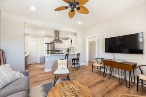 A modern living room with a sofa, coffee table, and dining area. A kitchen is visible in the background with a large TV mounted on the wall. - Coronado Unit 1 - B Ave Bungalows · King-sized 1/1, 5-10 Min Walk to Coronado Beach! (Coronado)