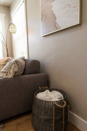 Eine Ecke eines Raumes mit einem grauen Sofa, einem Korbwäscheschrank mit weißen Gegenständen und einem gerahmten Kunstwerk an der Wand.