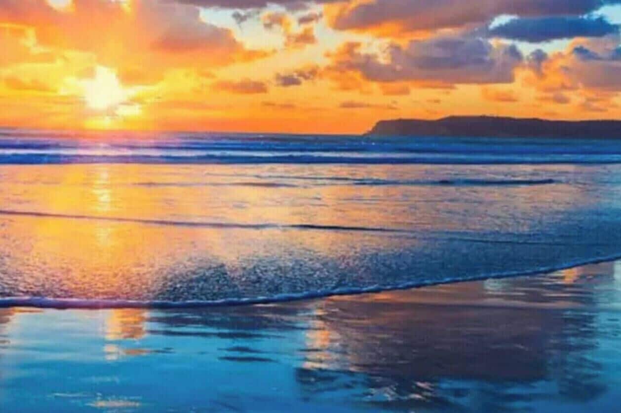 Ein Strand bei Sonnenuntergang, ruhiges Wasser und eine entfernte Küstenlinie.