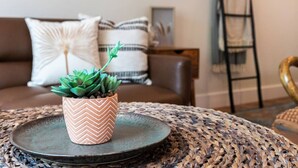 A living room with a brown sofa, a patterned cushion, a small potted plant, and a woven table. - Back Building A-C - B Ave Bungalows · All 3 Studios, 5-10 Min Walk to Coronado Beach! (Coronado)