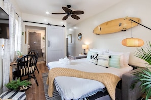 A hotel room with a bed, a ceiling fan, a surfboard, and a dining area with chairs.