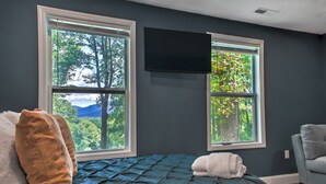 Une chambre à coucher avec un lit, deux fenêtres, un téléviseur fixé au mur et une vue sur les arbres et les montagnes.