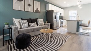 Un salon moderne avec un canapé gris, des coussins à motifs noir et blanc, un pouf noir et un tapis rayé. On aperçoit également un coin cuisine avec un réfrigérateur, un four à micro-ondes et un évier.