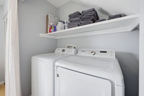 A laundry room with two front-loading washing machines and a shelf above holding folded towels and laundry supplies.