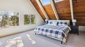Un dormitorio con techo inclinado, una cama con colcha de cuadros, dos mesitas de noche con lámparas y ventanas amplias que dan a vistas de árboles.
