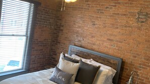 Quarto com parede de tijolos, cabeceira de madeira, cama com roupa de cama branca e várias almofadas, ventilador de teto e janela com persianas.