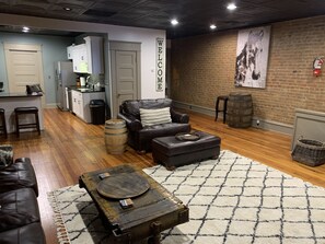 A modern living room with a leather sofa, a coffee table, and a rustic barrel. A kitchen is visible in the background, and a 'WELCOME' sign is on the wall. - NEW “Bourbon on the Square” Downtown Rustic Loft (Elizabethtown)