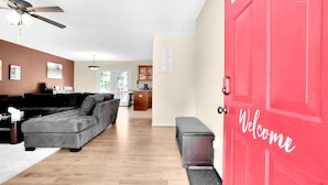 A living room with a grey sectional sofa, a wooden coffee table, and a ceiling fan. A red door with "Welcome" written on it is visible.