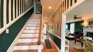 A staircase with wooden steps and white railings, leading to a living room with a fireplace, leather armchair, and a ceiling fan. - Marsa's Place-Marsh/Creek/Ocean Views; Easy Beach Access;Dogs Welcome (Edisto Island)
