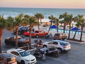 A parking lot with various vehicles, including cars and motorcycles, near a pool area with umbrellas and palm trees. - OCEAN FRONT STUDIO with 2 full beds & kitchenette (Daytona Beach)
