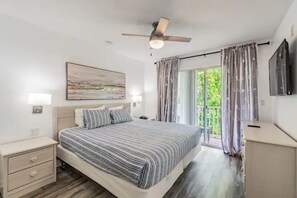 Un dormitorio con cama, mesitas de noche, ventilador de techo, un cuadro, televisor y balcón con cortinas.
