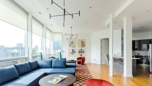 Un salon moderne doté d’une grande fenêtre, d’un canapé, d’un fauteuil rouge, d’une table basse et d’un espace cuisine visible à travers une porte ouverte.