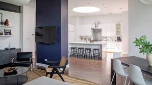 Un salon moderne doté d’un téléviseur à écran plat, d’un coin repas avec une table et des chaises, d’une cuisine avec des meubles blancs et un îlot central, et d’un grand luminaire suspendu blanc.