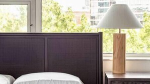 Une chambre d’hôtel avec un lit, une fenêtre donnant sur des immeubles et des arbres, une lampe sur une table de chevet et un meuble de rangement avec des tiroirs.