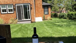Una mesa de madera con una botella de vino y dos copas, colocada al aire libre con una casa y un jardín al fondo.