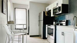 Una cocina moderna con armarios blancos, un refrigerador de acero inoxidable, un microondas, un horno y un lavavajillas. Hay un taburete de bar y una ventana con persianas.