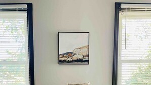 Una habitación con dos sillas blancas y una mesita, un cuadro en la pared y ventanas con persianas.