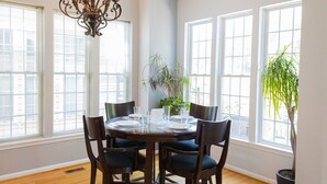 Un coin repas avec une table ronde dressée pour quatre personnes, entourée de chaises. De grandes fenêtres se trouvent d’un côté et une plante en pot de l’autre.