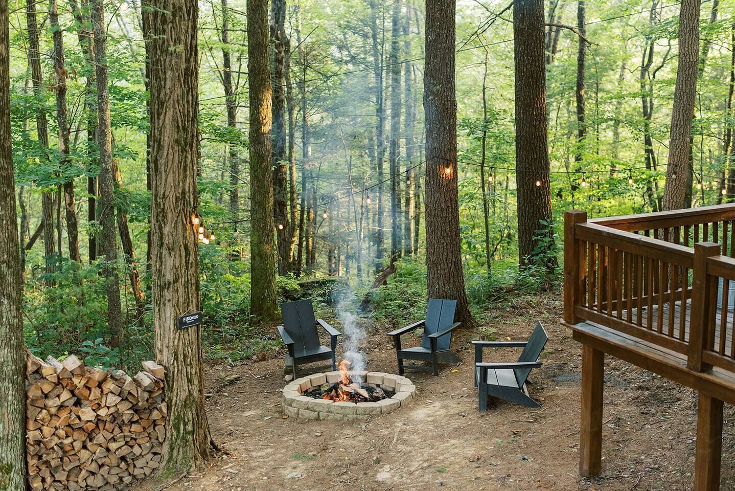 A forest setting with a fire pit, two chairs, and a wooden deck.