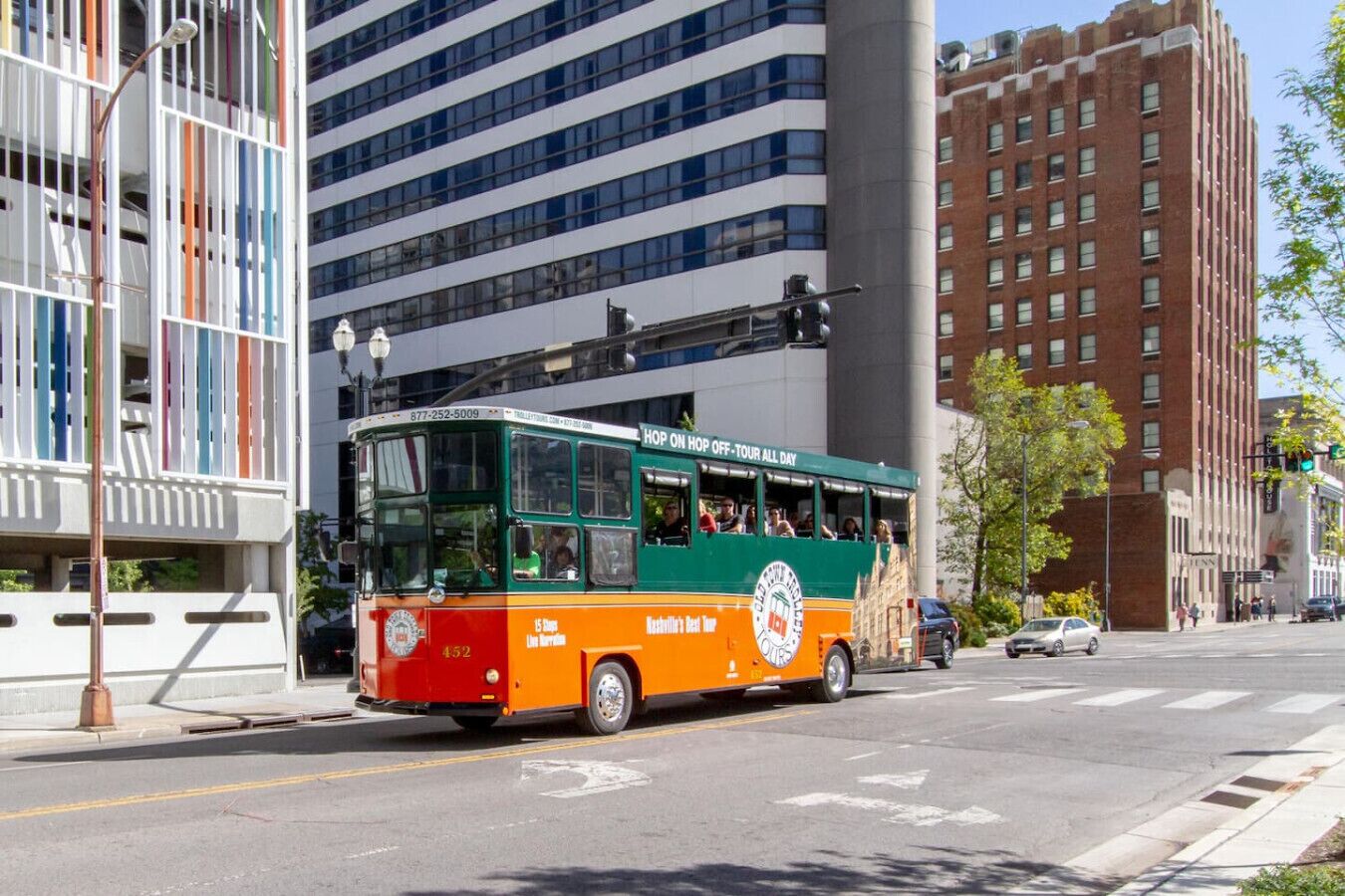 Un autobús verde y naranja con la leyenda "Hop on Hop off - Tour All Day" en la carrocería, circulando por una calle de la ciudad.