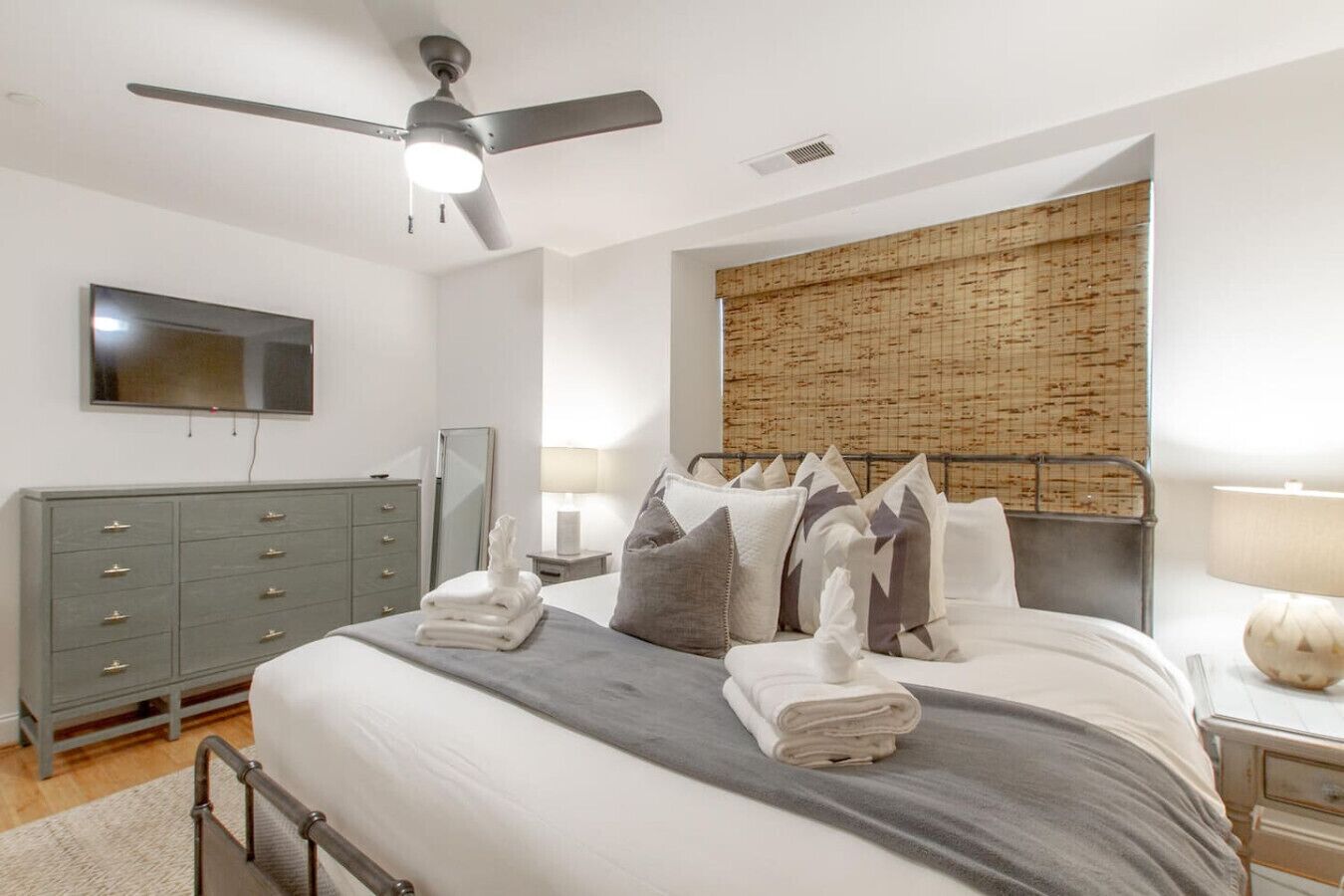 Un dormitorio con cama, ventilador de techo, televisor, cómoda y ventana con persiana de bambú.