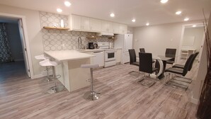 A modern kitchen with white cabinets, a central island with bar stools, a dining table with chairs, and a hexagonal patterned backsplash. - Beautiful two bedroom + den lower level suit (Calgary)
