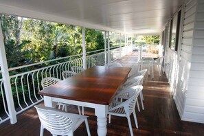 Ein überdachter Balkon mit Holztisch und weißen Stühlen, umgeben von einem weißen Geländer, mit Blick auf einen üppigen Garten.
