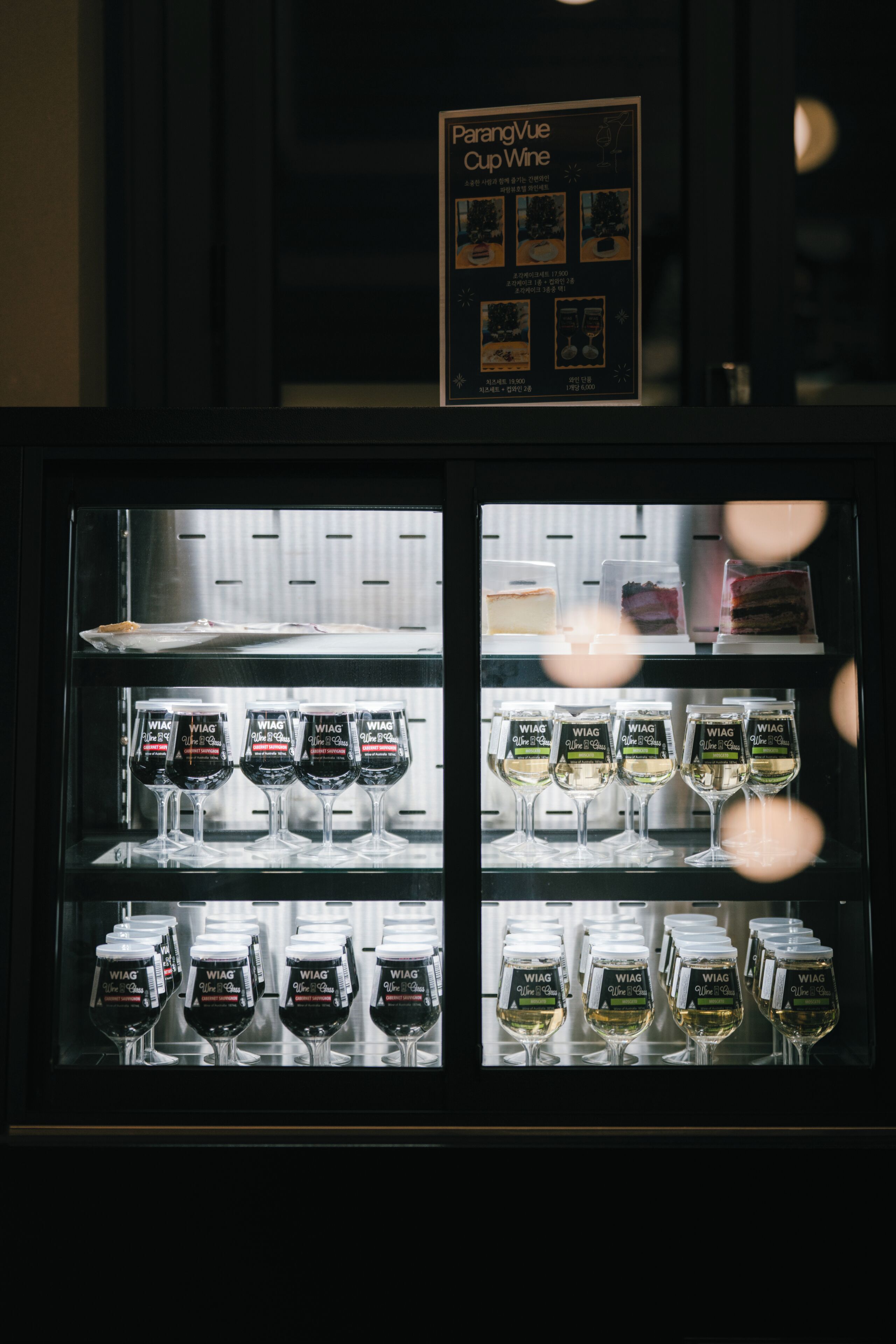 A refrigerated display case with wine glasses and bottles, a menu sign above, and a reflection of lights.