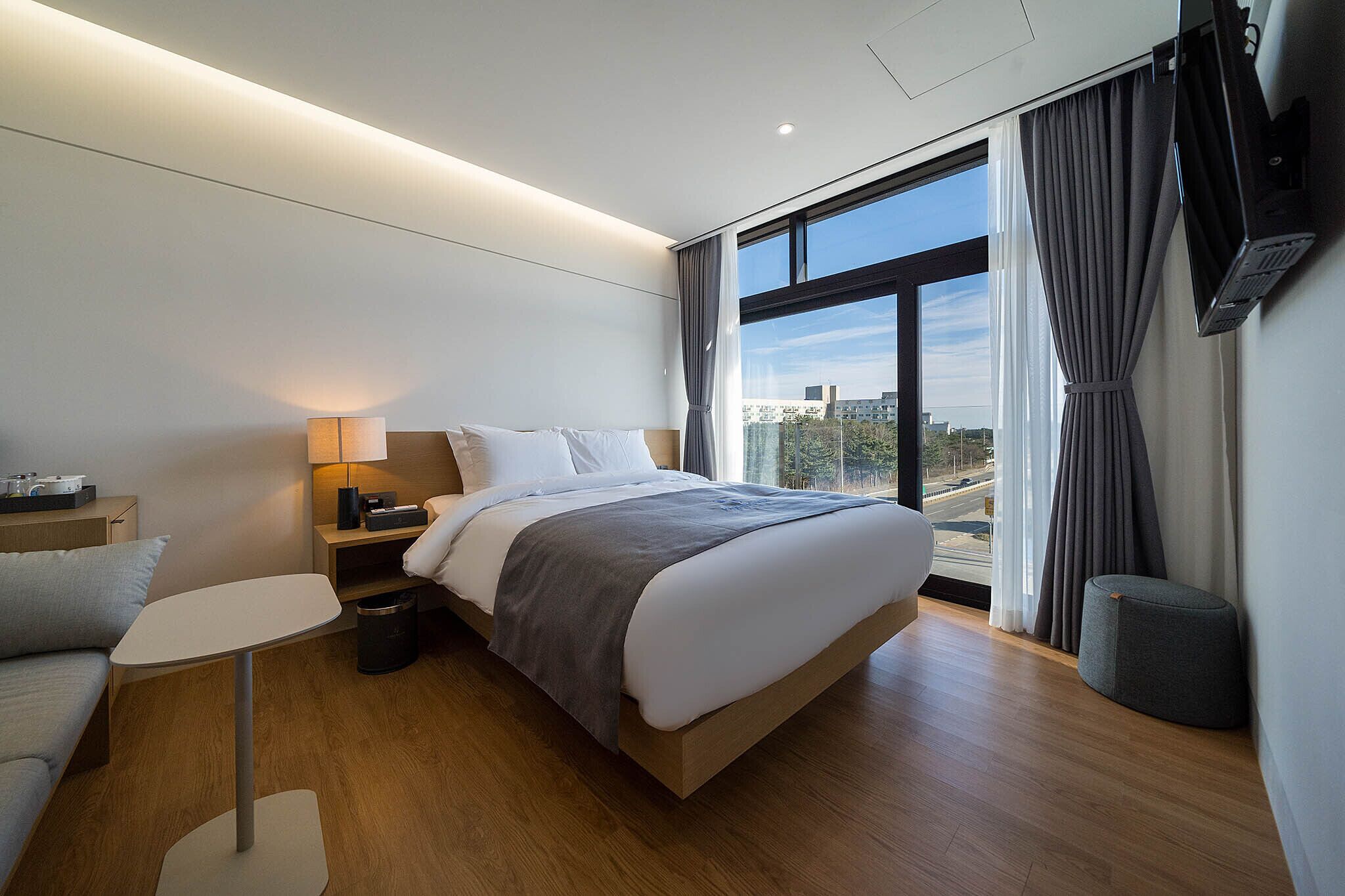 A modern hotel room with a large bed, a small table, a sofa, and a TV mounted on the wall.